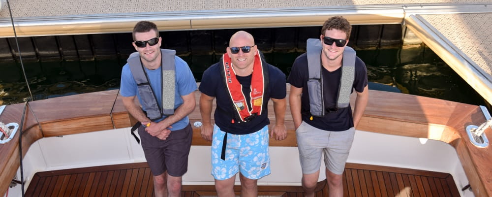 three men wearing lifejackets standing on the back of a boat looking up at camera