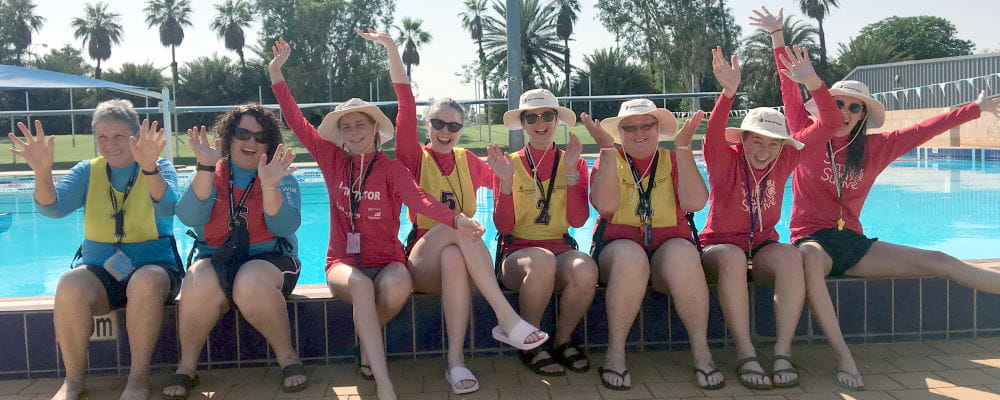 Eight swim instructors sitting by the pool with arms in the air at South Hedland Aquatic Centre
