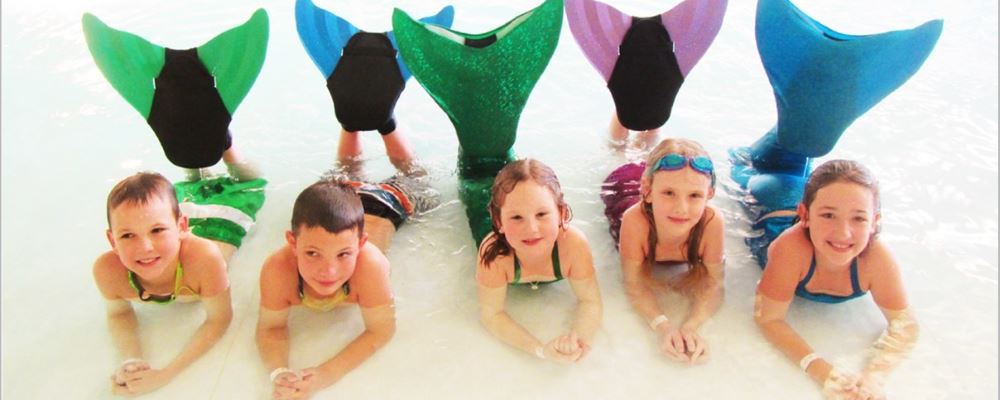 two boys and 3 girls wearing mermaid tails and monofins laying in the water smiling at camera