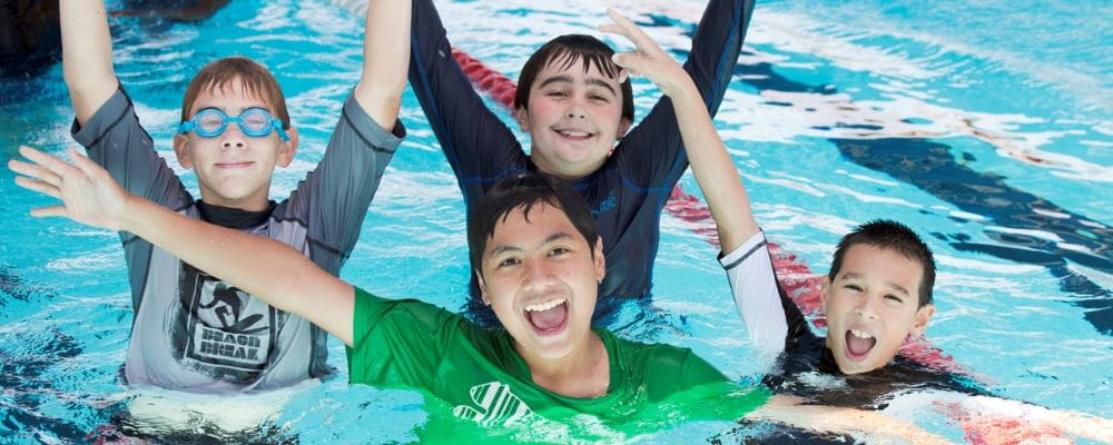 Four boys in the pool with arms in the air, smiling