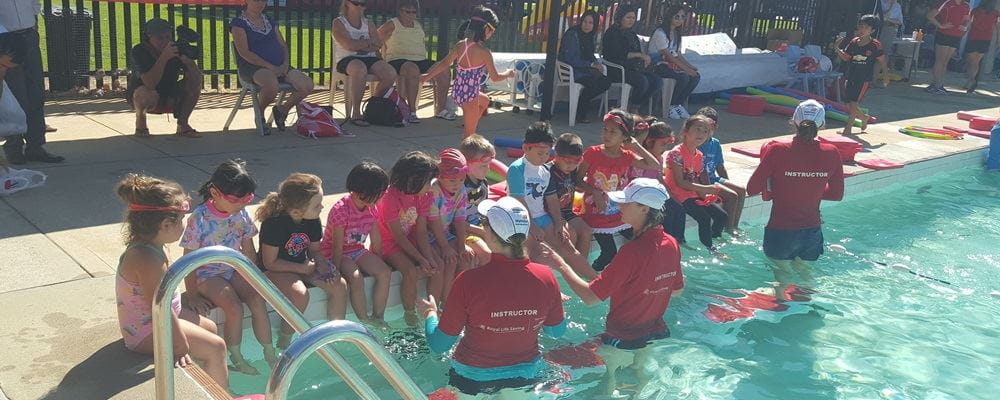 multicultural children sitting on the edge of the pool with 3 instructors ready to start a lesson