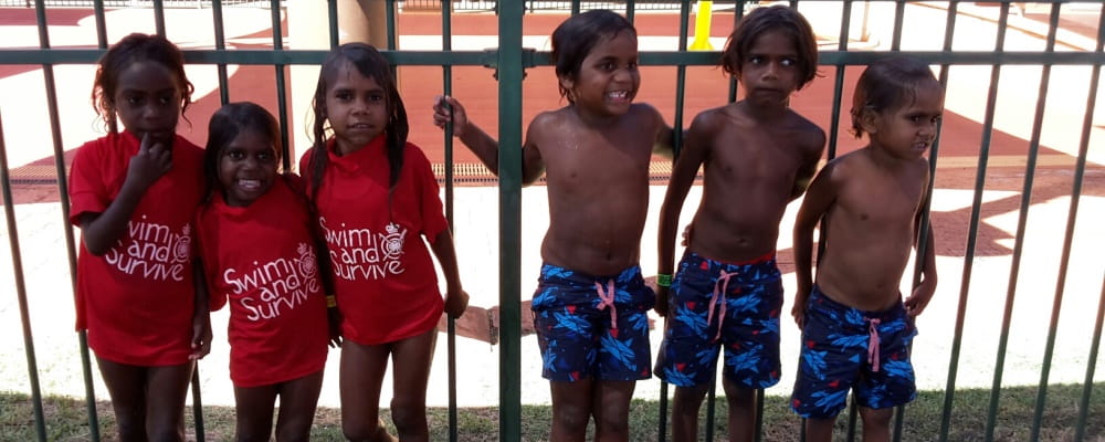 aboriginal children wearing swim and survive rashies and board shorts by a pool fence
