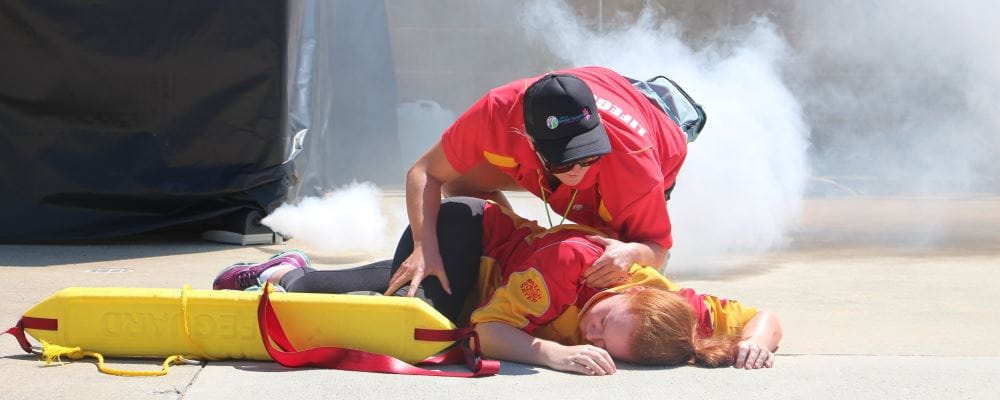 A lifeguard attending to a person passed out on the ground with smoke in the background