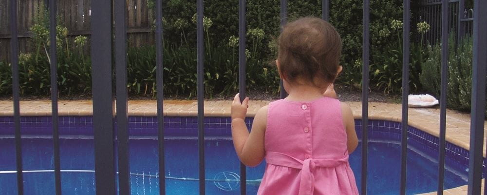 image of toddler girl holding on to pool gate and looking at pool