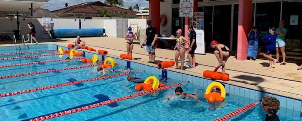 Children lining up along the side of a pool while other kids are in the water starting a manikin tow race