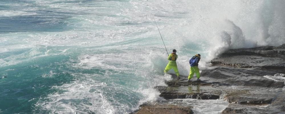 Two men fishing from rocks with a large waves crashing