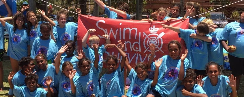 Children wearing blue Spirit Carnival t-shirts, standing in front of a Swim and Survive banner