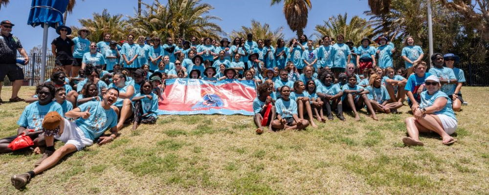 Group photo of 100 plus Aboriginal children who participated in the spirit carnival. Wearing light blue shirts with design of seagrass and turtles swimming. 