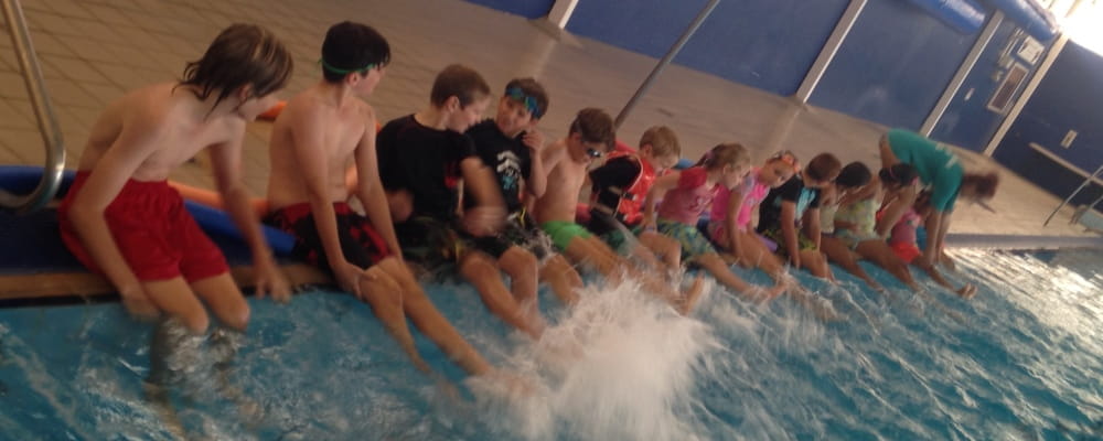 Children sitting lined up on the side of a swimming pool kicking their legs and splashing