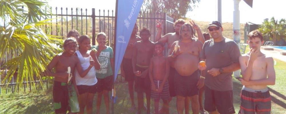 A group of children enjoying eating their fruit standing with Marble Bar Swimming Pool Senior Aquatic Officer, David Clarke