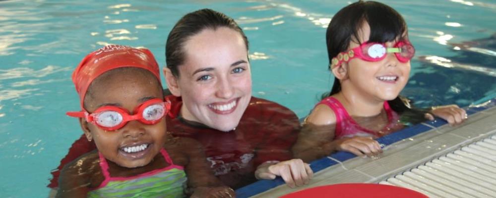 image of two young girls with a swimming instructor at the edge of the pool smiling at the camera