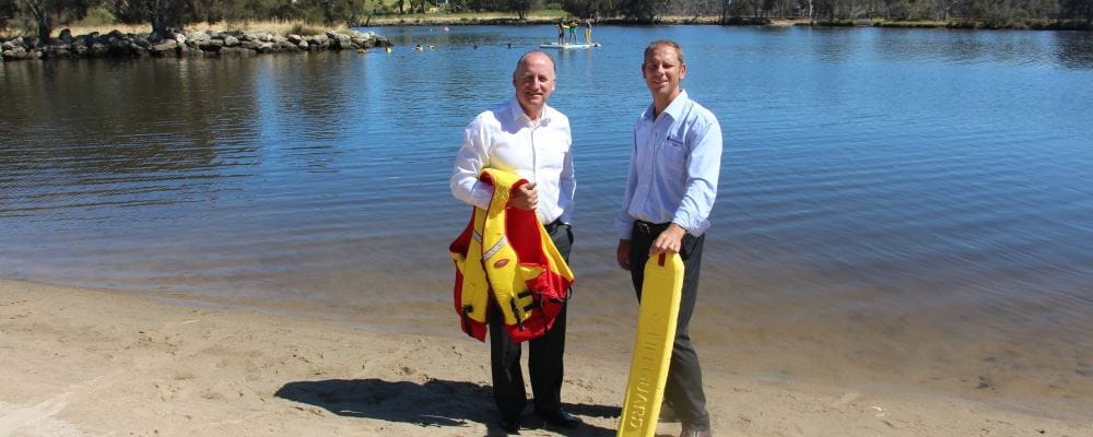 Federal Member for Swan Steve Irons holding a lifejacket, with RLSSWA CEO Peter Leaversuch holding a rescue tube, standing by the Swan River with children learning rescue skills in the background"