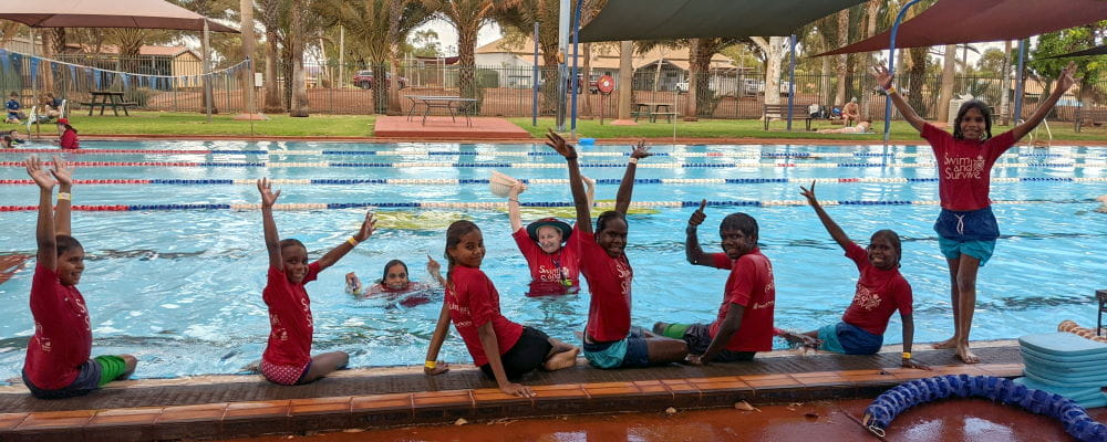 Aboriginal children along the edge of a pool with their instructor in the water, all with arms in the air