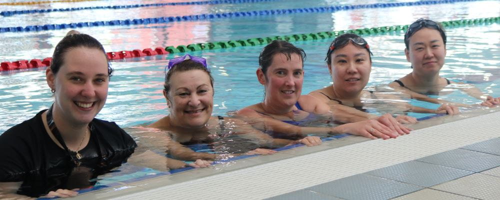 A swim instructor with four women standing in a pool, holding onto the edge