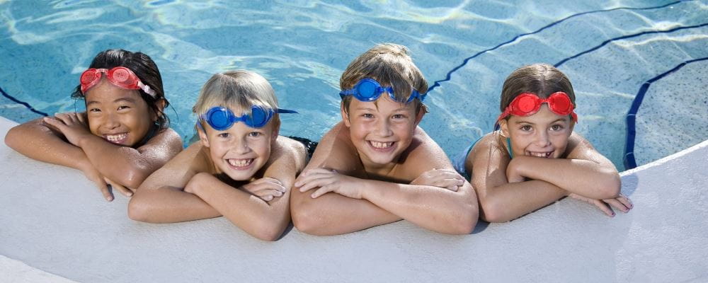 image of four children leaning on the edge of a pool wearing goggles and smiling at the camera
