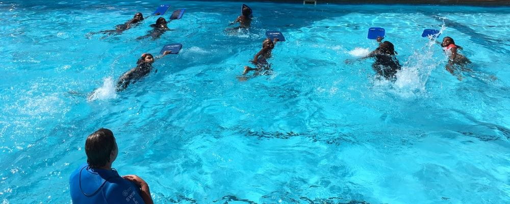 Image of aboriginal children swimming with kickboards across a pool, with teaching looking on