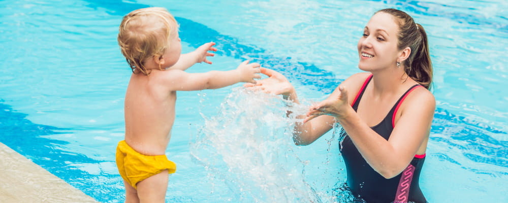 A baby standing on the pool edge with mum facing him in the water splashing