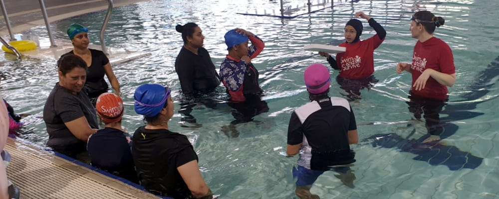 A group of women in the pool with two swim instructors explaining the class