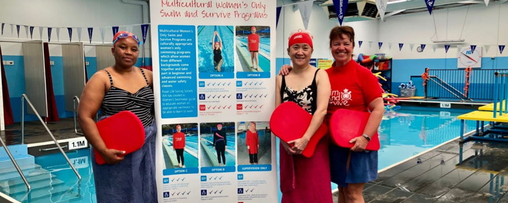 Two multicultural women with their instructor standing by the pool with a women's only swimming lessons sign