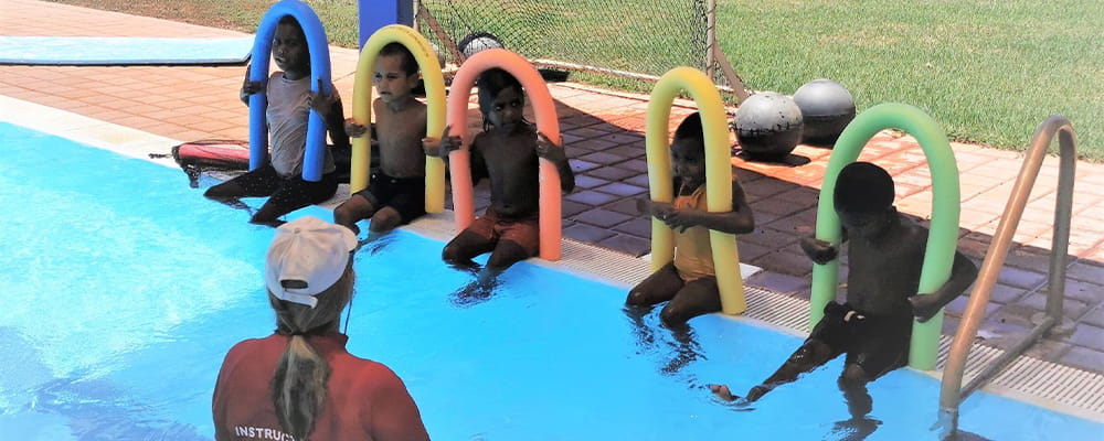 Aboriginal children sitting along the edge of a pool with pool noddles and their instructor in the water
