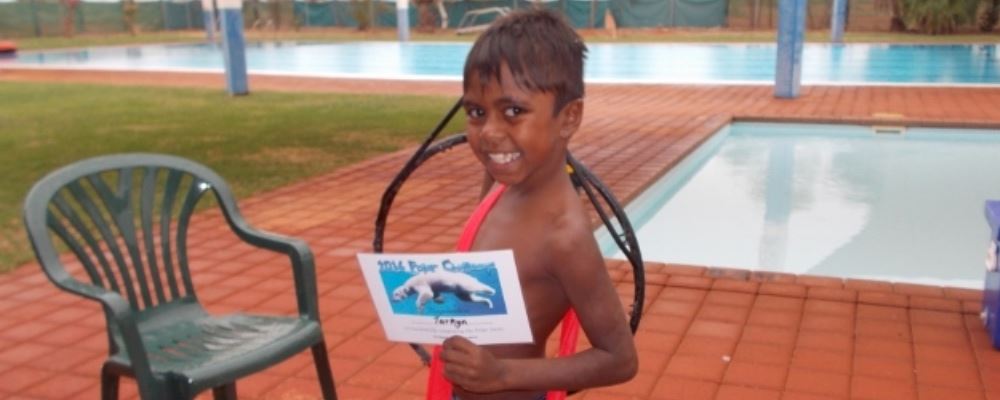 a Yandeyarra boy standing by the pool with his swim for fruit certificate, smiling at the camera