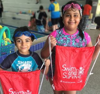 Two multicultural children by the pool at Bayswater Waves, holding Swim and Survive bags