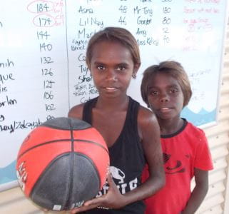 Two aboriginal boys with a basketball in front of the Go For 2 and 5 Tally Board at Bidyadanga