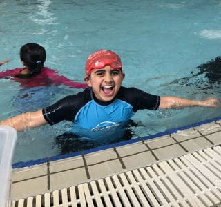 An islamic boy in the pool wearing a swimming cap and goggles, with arms out to the side smiling