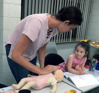 A mum doing CPR on a baby manikin with her toddler daughter looking on
