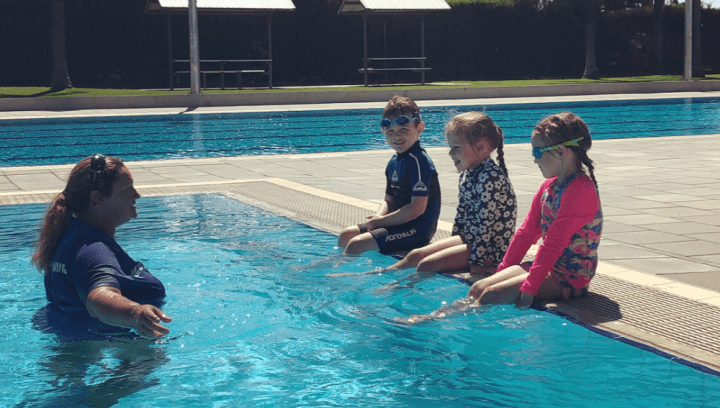A swim instructor in the pool with three children sitting along the edge