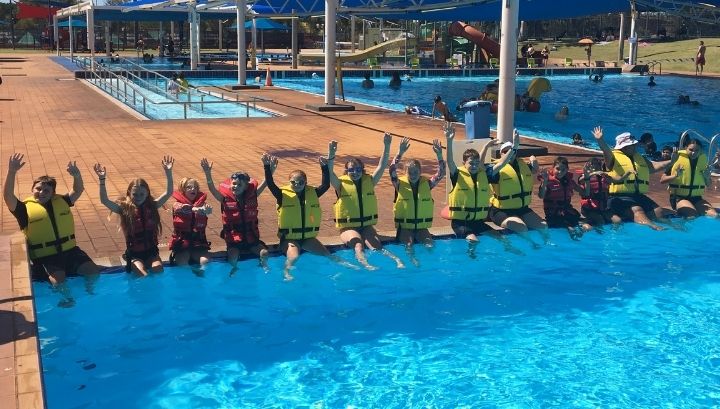 Children and police sitting along the edge of the pool wearing lifejackets