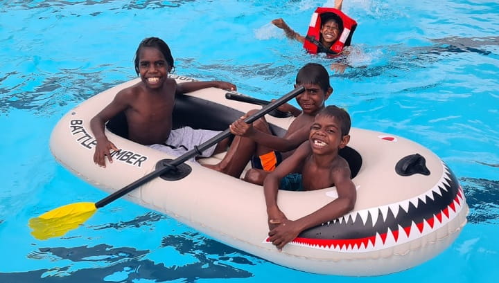 three Aboriginal boys smiling in an inflatable boat in a pool