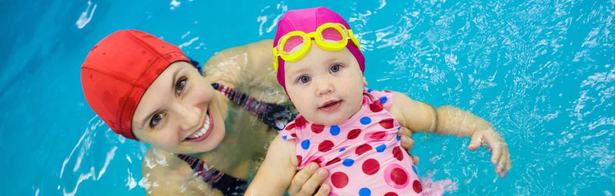 Image of mum with red swim cap on holding baby girl in spotted bathers, pink swim cap and yellow goggles floating on back in a pool