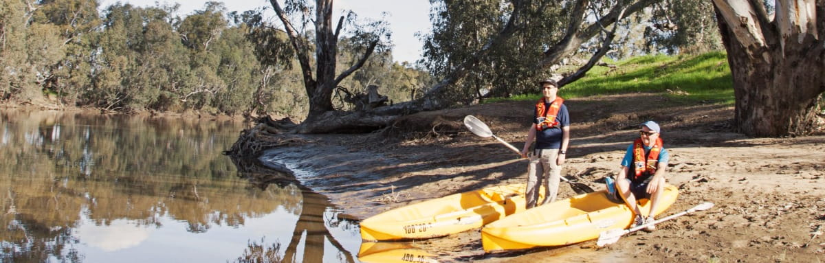 Two men wearing lifejackets and holding paddles are standing beside two yellow kayaks on a riverbank