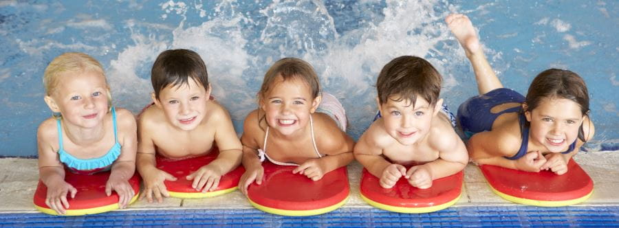 Five children leaning on the edge of the pool with red kickboards, smiling at the camera
