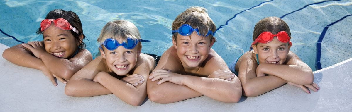 Image of 4 children leaning against the edge of a pool, wearing goggles on their head and smiling at the camera