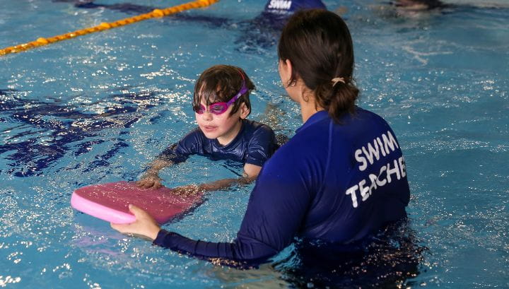 Female swim teacher wearing long sleeve blue swim shirt with "Swim Teacher" written on the back. Helping a young boy swim with a red kick board. 