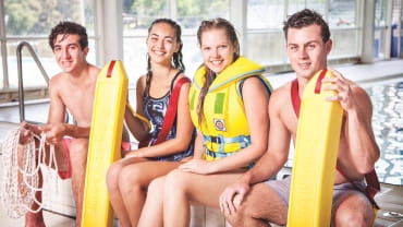 Four young adults sit on the side of an indoor swimming pool; one is wearing a lifejacket, two are holding rescue tubes and one is holding a length of rope