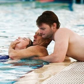 Image of a man leaning over edge of pool helping another man who is assisting a female patient