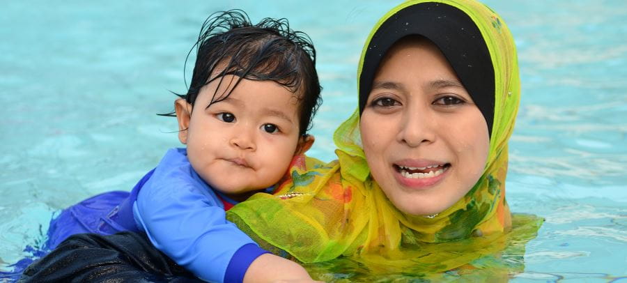 A woman with a Muslim headdress on in the pool with her baby on her back