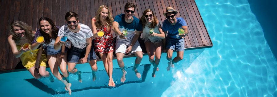 A group of young people sitting along the edge of a deck holding alcoholic drinks with legs dangling into a swimming pool
