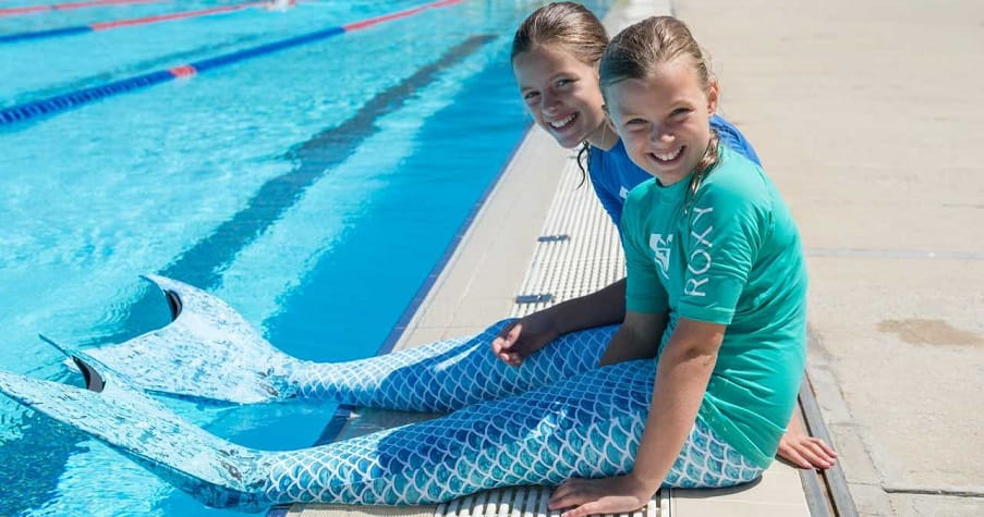 Two girls wearing blue mermaid tails sitting on the edge of a pool