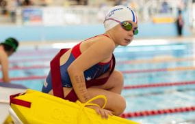 A young girl wearing goggles and holding a lifeguard rescue tube, crouching on the blocks by the pool