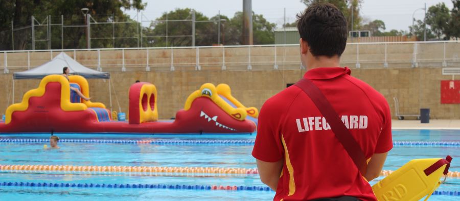 A lifeguard with back to the camera wearing a red shirt and holding a rescue tube, standing by the pool with a large pool inflatable in the distance