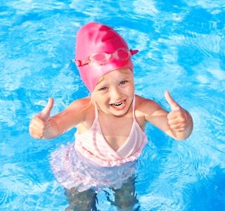 Image of young girl wearing pink swim cap, bathers and goggles giving the two thumbs up sign while in the pool