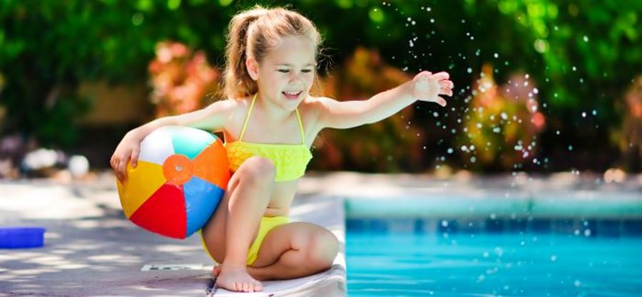 A young girl by the pool holding a beach ball and splashing the water