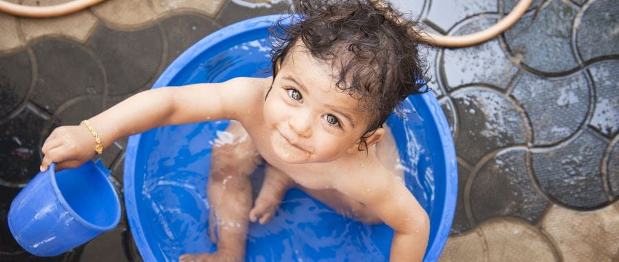 A toddler sitting in a blue bucket full of water, holding a smaller blue bucket in his hand and looking up at the camera