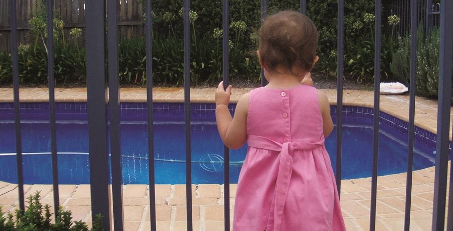 A toddler girl wearing a pink dress standing by a black metal pool fence