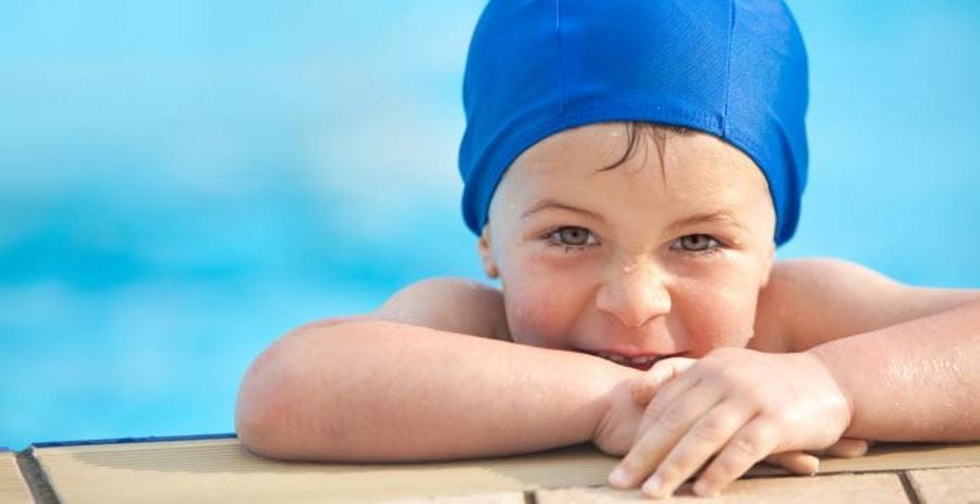 A little boy leaning on the edge of a pool wearing a blue swimming cap