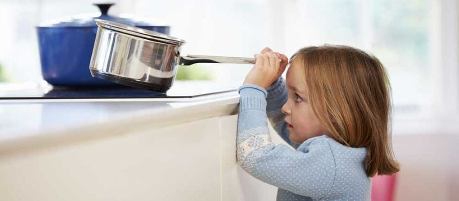 A toddler girl holding the handle of a pot and just about to pull it off the stove